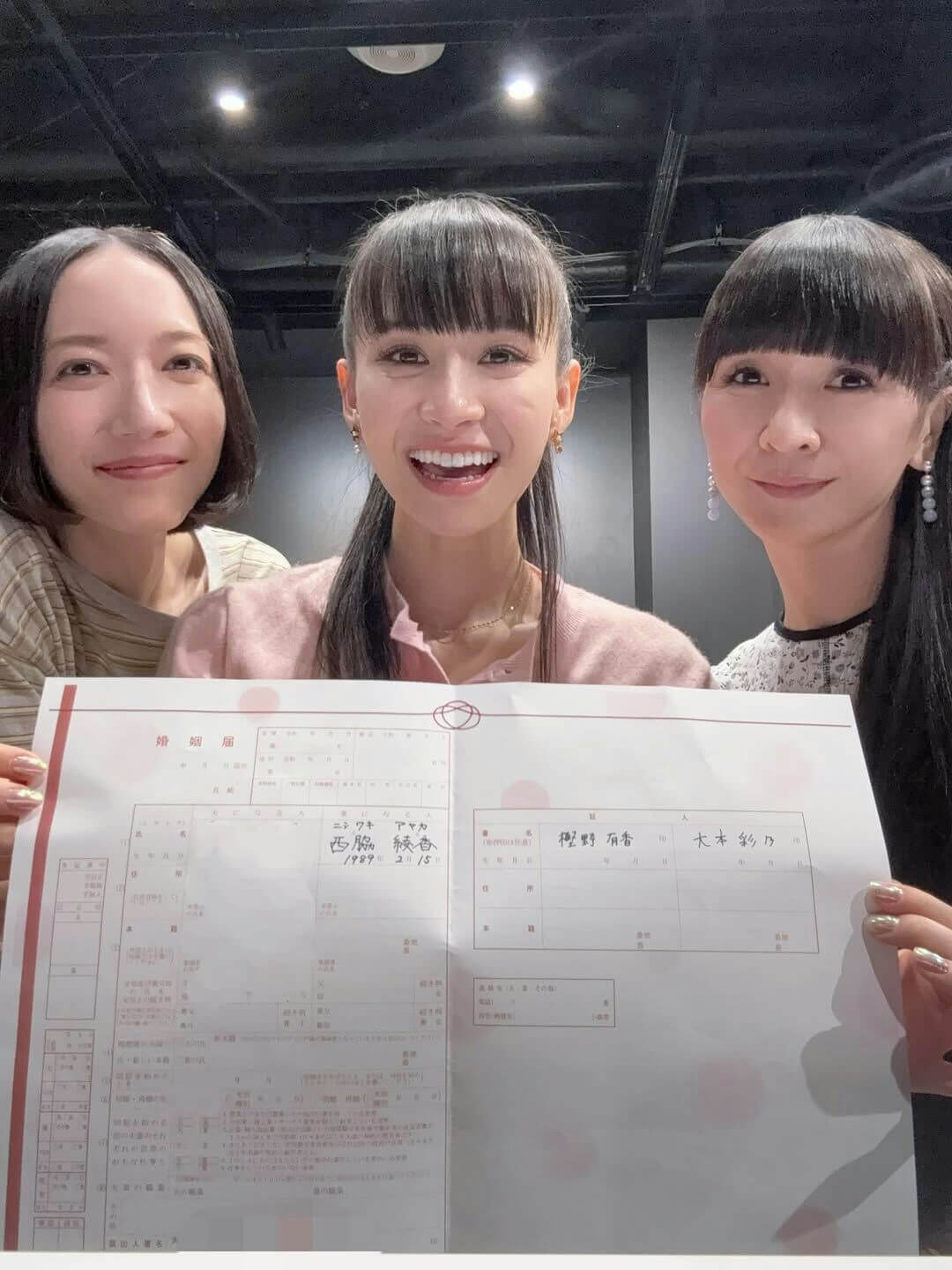 people-three women smiling-A-holding document-A-Japanese marriage registration form (kon'in todoke)-B-indoors with spotlights-C-celebratory atmosphere