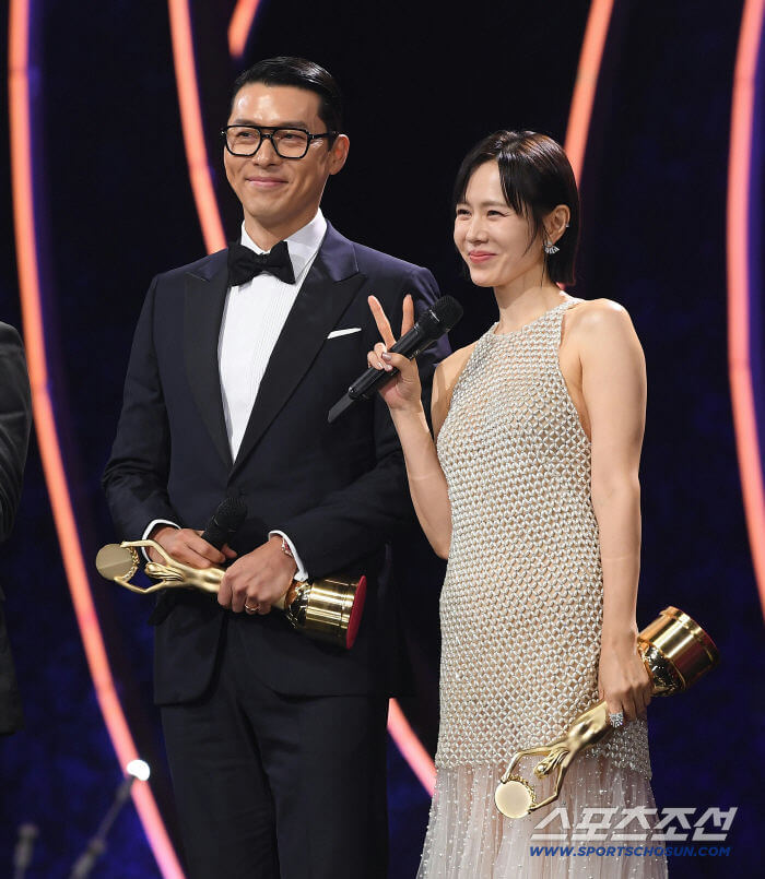 people-Two-A-Korean-celebrities-A-man-in-a-tuxedo-and-glasses-A-woman-in-a-sparkling-dress-holding-a-microphone-and-making-a-peace-sign-Both-holding-gold-trophies