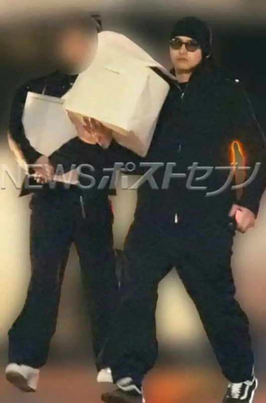 people-two individuals-A-dressed in black attire-A-one person hiding face with large white item-B-one person wearing sunglasses and a beanie-C-Japanese text overlay "NEWSポストセブン"