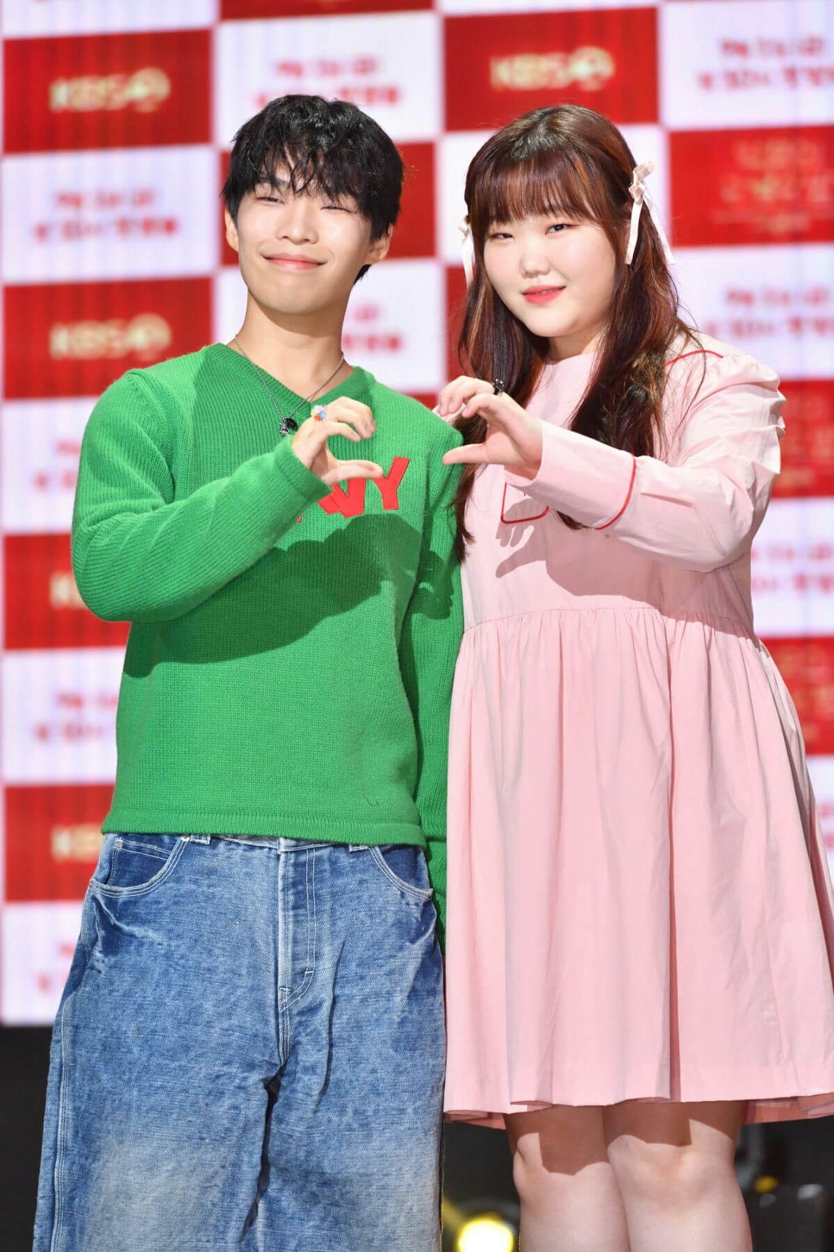 people-male and female-hand heart gesture-male wearing green sweater and jeans-female wearing pink dress-red and white checkered background