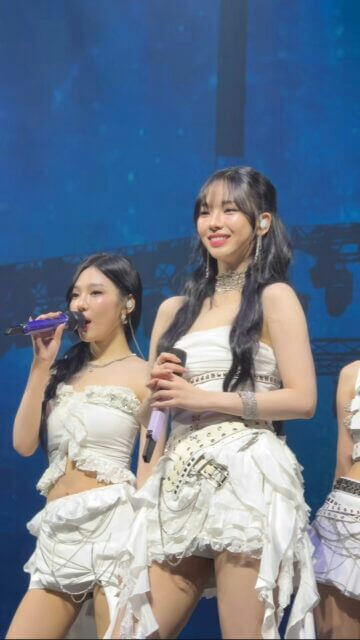 people-Two young women-A-performing on stage-A-wearing white outfits-A-holding microphones-B-smiling-C-long dark hair