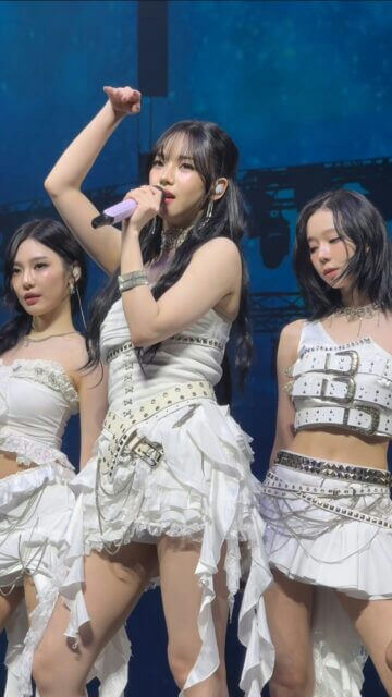 people-three women-A-kpop-A-stage performance-A-white outfits-A-singing-A-microphone-B-black hair-B-straps and buckles-C-ruffled skirts