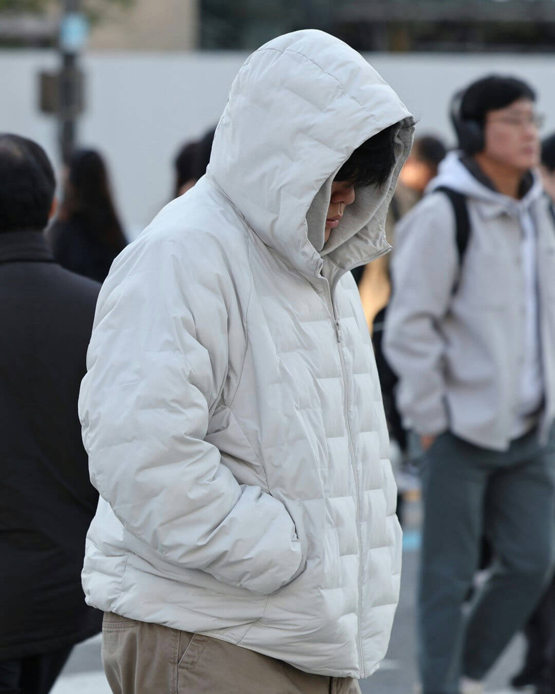 people-person in light gray puffer jacket with hood up-person in background wearing headphones-outdoor setting-casual attire