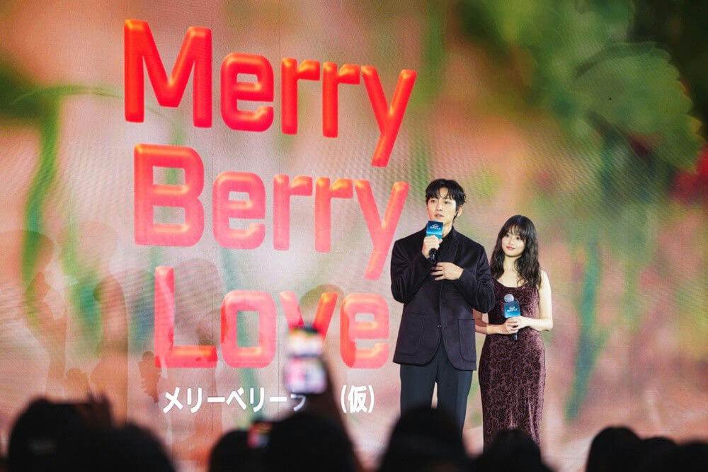 people-Two people (a man and a woman)-A-speaking into a microphone-A-standing on stage-A-promotional event-B-large screen display-B-red 3D text
