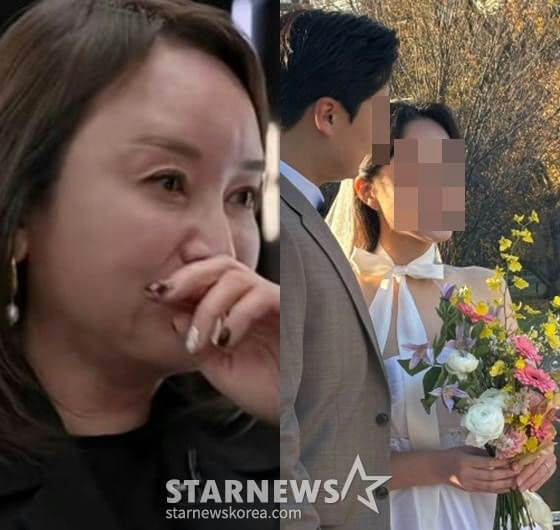 people-woman crying while covering mouth-wedding couple posing outside with flowers-woman with brown hair and pearl earring-man in suit next to bride-bride in white dress with bow and veil-colorful bouquet-STARNEWS watermark