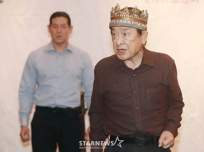 people-One older man-wearing a crown-One younger man-standing in background-security/bodyguard-brown shirt-light blue shirt