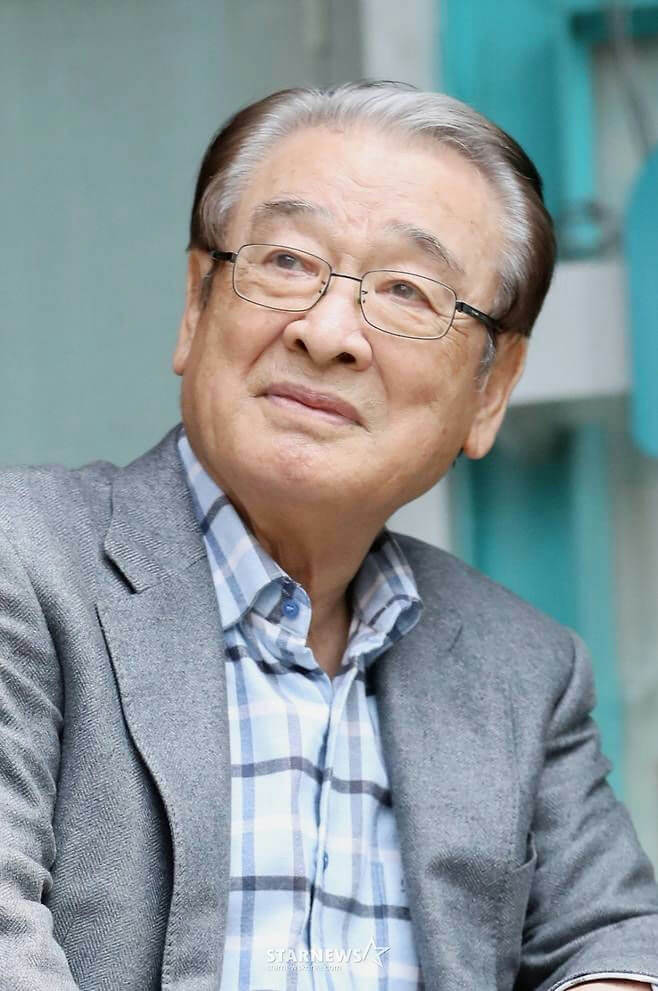people-An elderly East Asian man-A-wearing glasses-A-with graying hair styled back-A-dressed in a gray tweed-like blazer over a light blue and white plaid shirt-A-looking slightly up and to the left