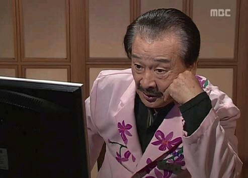 people-older man-pink floral robe-looking at monitor-hand on cheek-MBC logo