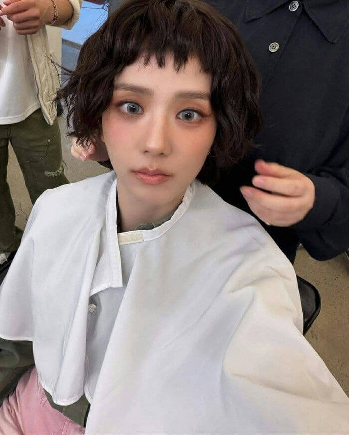 people-woman-A-short dark hair-A-micro bangs-A-heavy blush makeup-A-contact lenses-A-white salon cape-A-in preparation-A-looking at camera