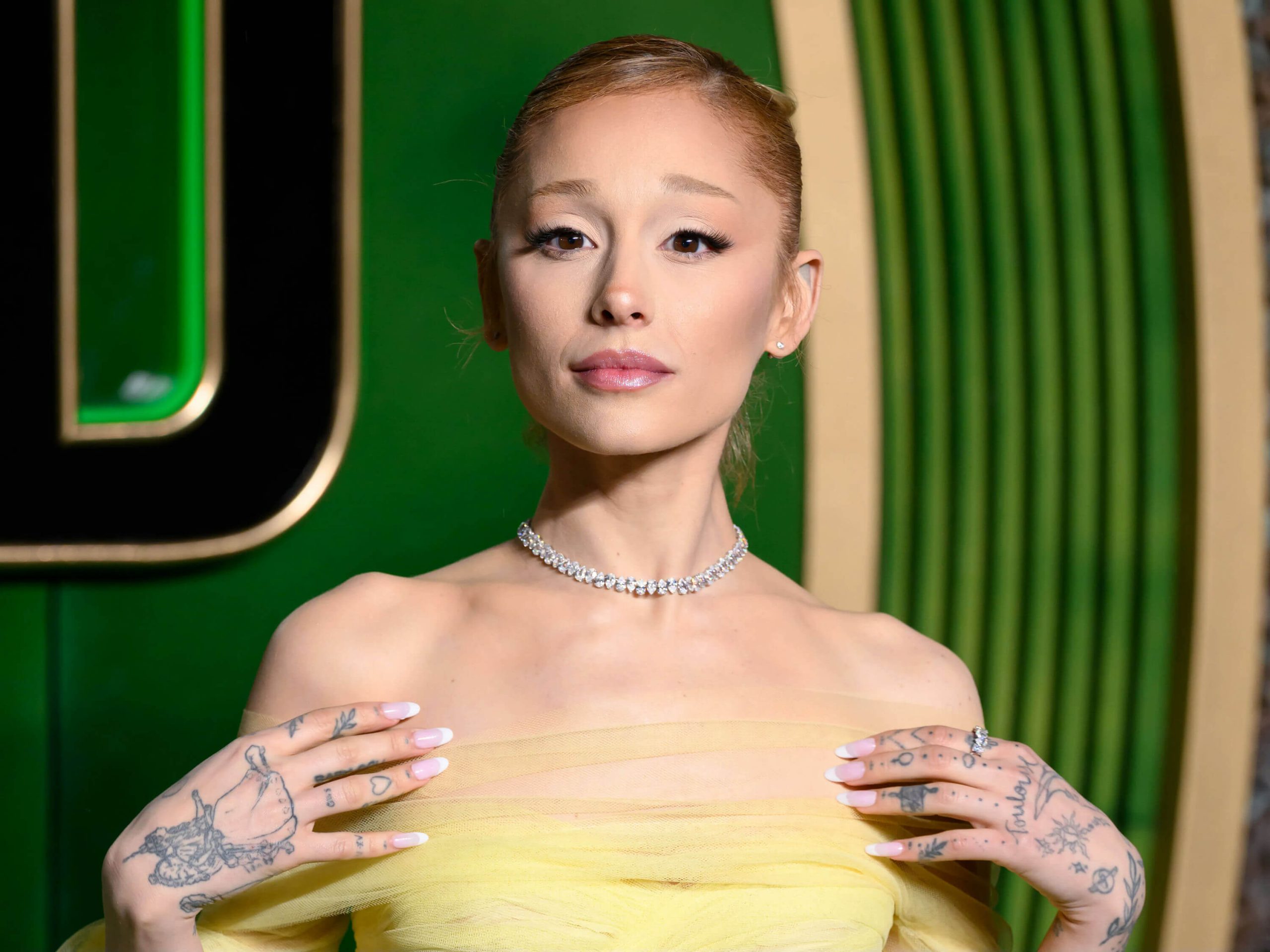 people-Ariana Grande-wearing yellow tulle gown-wearing diamond choker necklace-showing hand tattoos-posing indoors with green and black background