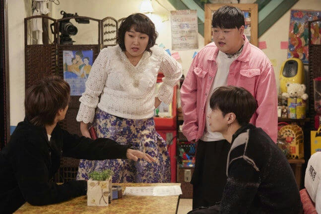 people-Four people-A-indoor setting-A-casual gathering-A-discussion-B-woman in white top and patterned skirt-B-man in pink jacket-C-man in black jacket gesturing-C-man in black sweater listening