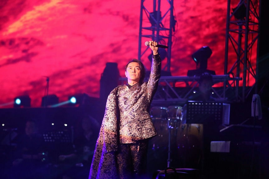 people-Man singing-A-Stage performance-A-Red background lighting-A-Traditional/brocade outfit-A-Microphone raised-A-Dark stage-A-Concert setting