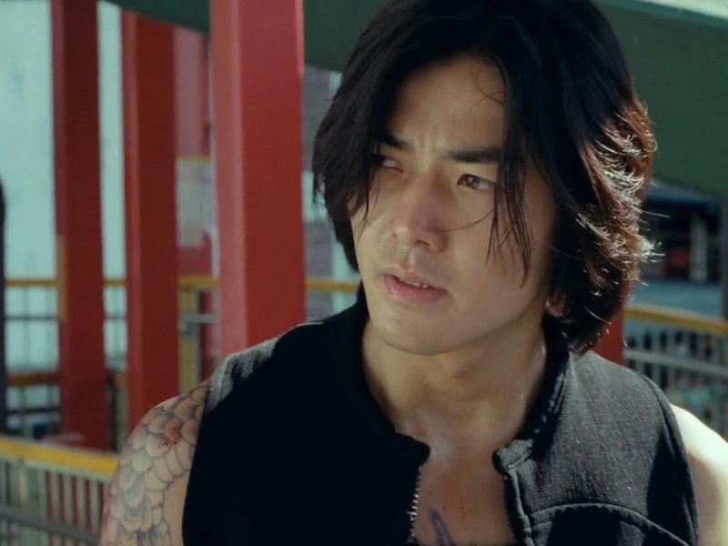 people-Asian male-A-long dark hair-A-black sleeveless jacket-A-tattooed arm-A-serious expression