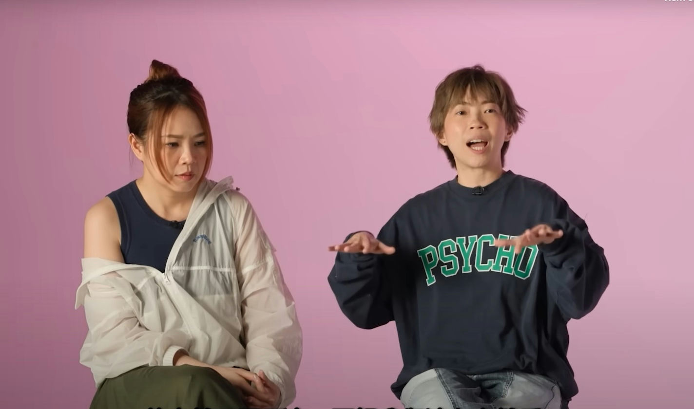 people-two women-sitting-pink background-woman on left wearing athletic gear and looking down-woman on right wearing dark sweatshirt with 
