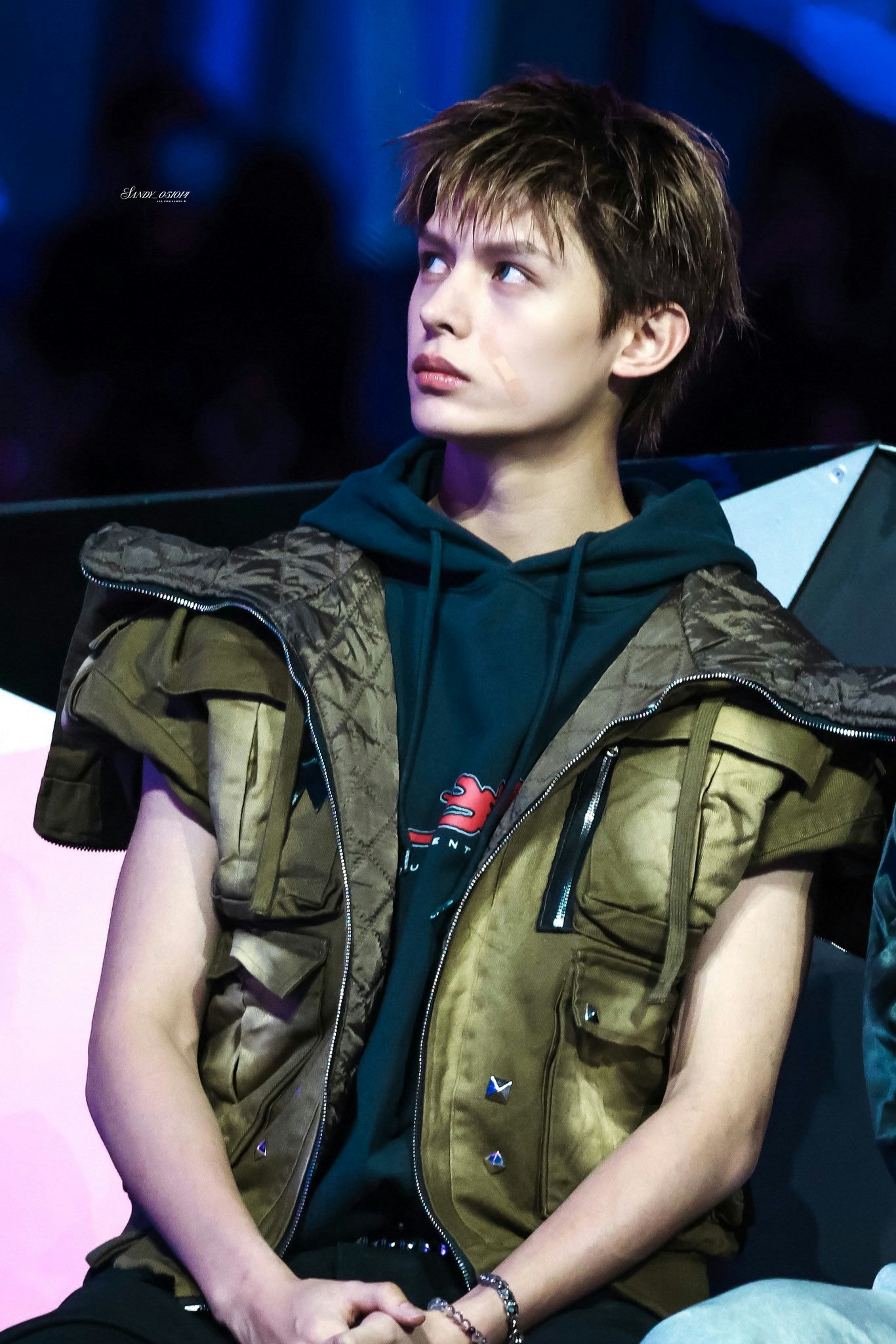 people-young man-A-looking up-A-brown shaggy hair-A-green hoodie-A-khaki quilted vest-A-light makeup scar