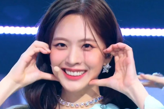 people-young woman-smiling brightly-A-making finger heart gesture-B-wearing sparkly choker necklace-C-on stage with blue lighting