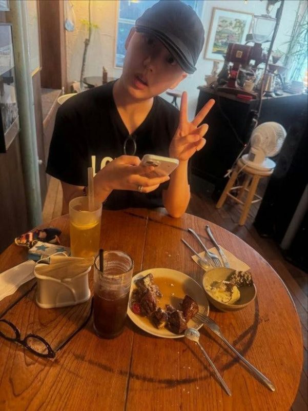 person-sitting at table-making peace sign-holding phone-wearing black t-shirt and black cap