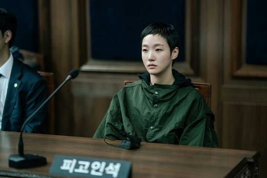 people-woman with short dark hair and a green jacket-A-in a courtroom or hearing setting-A-seated at a wooden desk-A-sign in front reads "피고인석" (Defendant's Seat)-B-male figure partially visible to the left in a suit-C