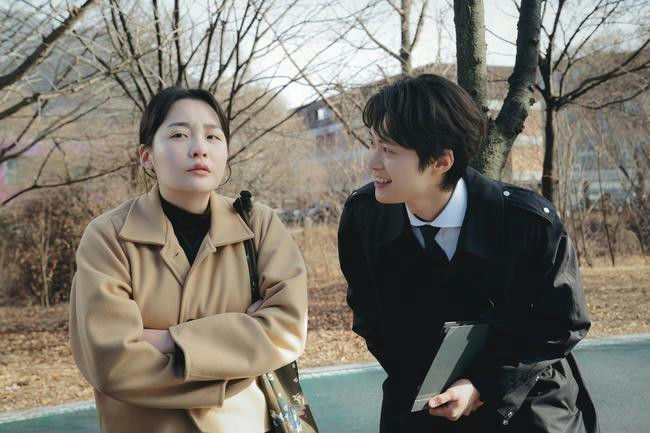 people-Woman-Serious expression-Man-Smiling and leaning in-Outdoor setting-Winter trees-Tan coat-Black coat