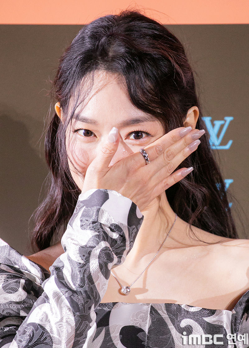 people-woman-covering mouth with hand-hand features ring and manicured nails-off-the-shoulder patterned top-long dark hair-Louis Vuitton logo visible in background