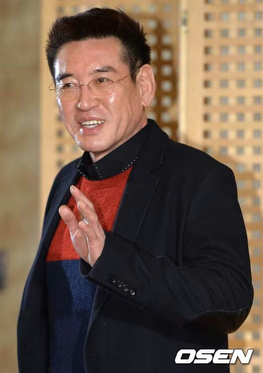 people-An older man-A smiling face-A hand waving-A black blazer-A color-block sweater