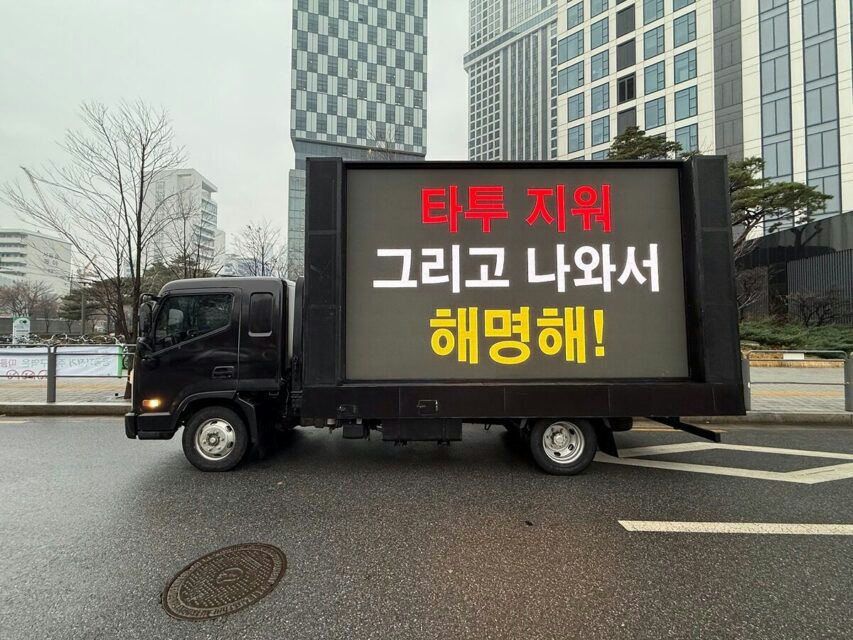 A black truck with a large LED screen attached to its cargo area is parked on a wet street in an urban setting, featuring tall modern buildings in the background-A-LED screen-A-Korean text-A-protest vehicle-A-city street