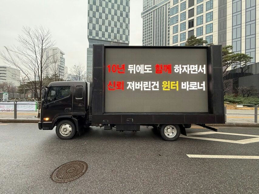 A dark-colored truck with a large LED screen is parked on a wet city street-A-digital billboard-A-Korean text-A-urban environment-A-broken promise