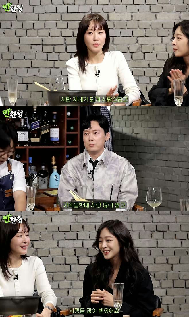 people-Woman in white top talking-Woman in black smiling-Man in patterned shirt listening-Setting in a dimly lit room with a brick wall background-Subtitles in Korean: 