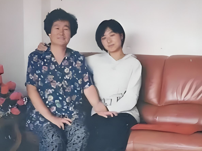people-Two individuals (likely two women)-sitting-A-red leather couch-A-white wall-B-floral print shirt-C-white hoodie