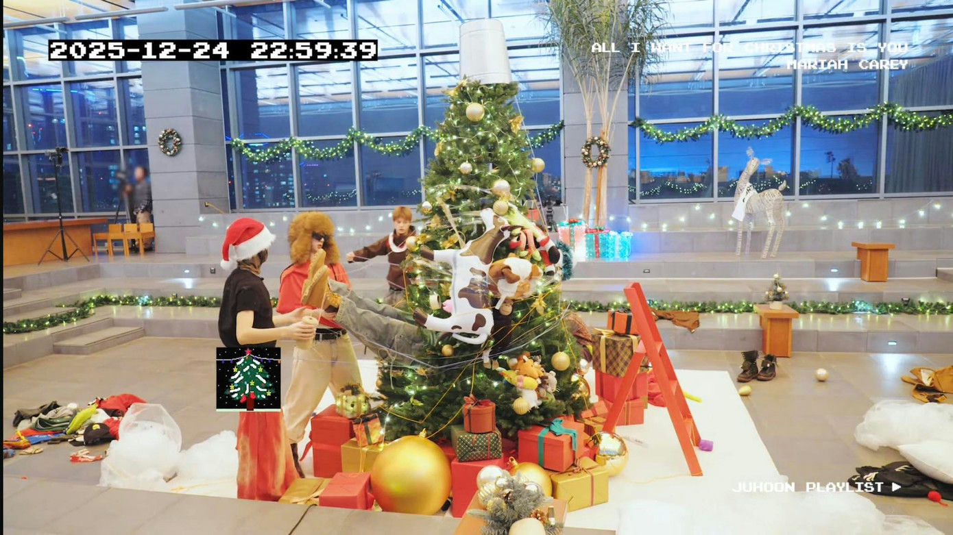 people-Two people in Santa hats and winter-style clothing decorating or interacting with a Christmas tree-A Christmas tree entangled with cow plush toys and lights-A bucket atop the tree-The setting appears to be a large, modern atrium or lobby decorated for Christmas with large windows in the background-A banner displays lyrics from Mariah Carey's 