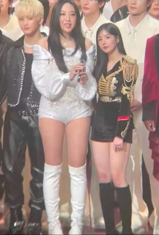 people-Two young women standing center-Two women wearing performance outfits-Woman on left wearing white off-shoulder romper and white thigh-high boots-Woman on right wearing black military-style jacket and black shorts with black knee-high boots-Blonde male figure partially visible on far left-Group of people standing in background