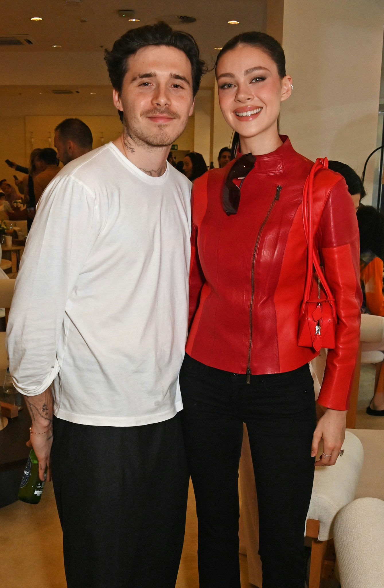 people-Brooklyn Beckham-white long-sleeve shirt-Nicola Peltz Beckham-red leather jacket-smiling-holding beer bottle