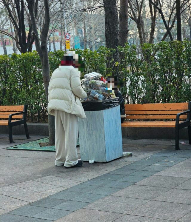 people-two figures-A-trash receptacle-A-outdoor setting-B-paved walkway-C-puffy coat