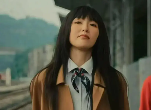 people-Asian woman-long black hair-bangs-brown coat-white collared shirt-black patterned neckerchief-outdoor setting-railway/station background