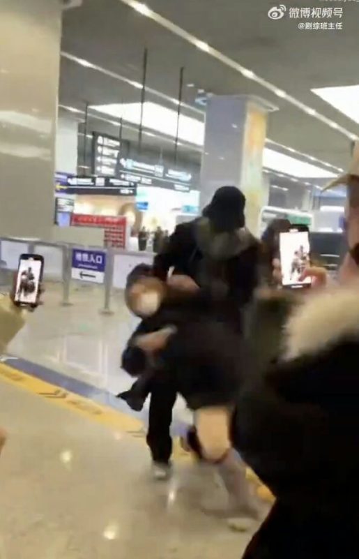 people-one adult figure carrying another smaller figure-carrying-airport/indoor setting-crowded/surrounded by phones-low light/motion blur