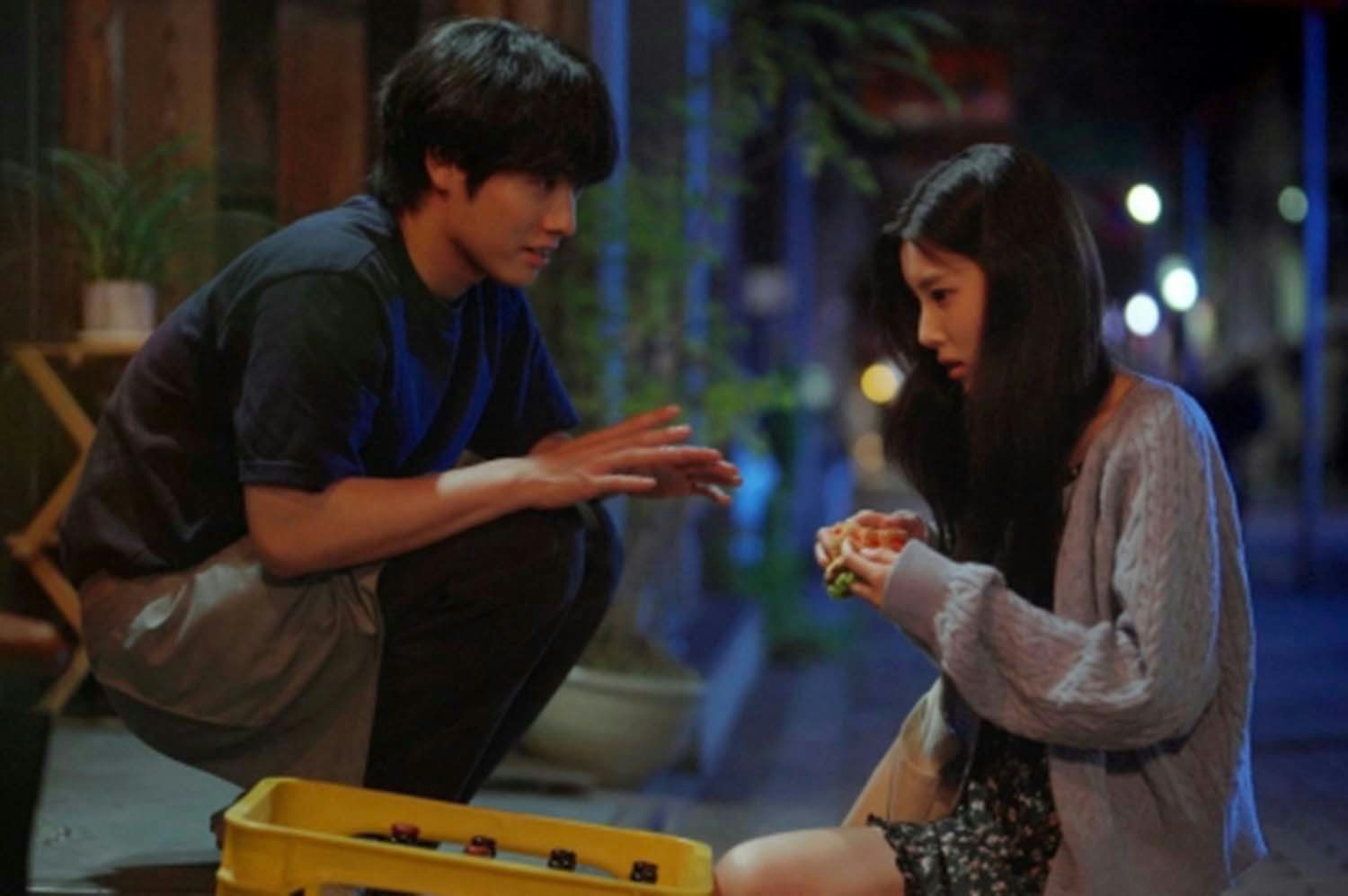 people-Two young adults (a male and a female)-interacting-outdoor setting at night-crouching male with dark hair wearing a dark blue shirt and light pants-female sitting wearing a gray cable-knit cardigan and holding food-yellow crate partially visible in the foreground