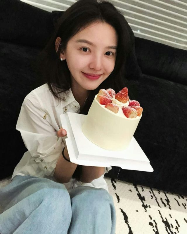 people-Kim So-eun-smiling-A-holding-a-small-strawberry-cake-A-sitting-on-a-rug-A-wearing-a-white-shirt