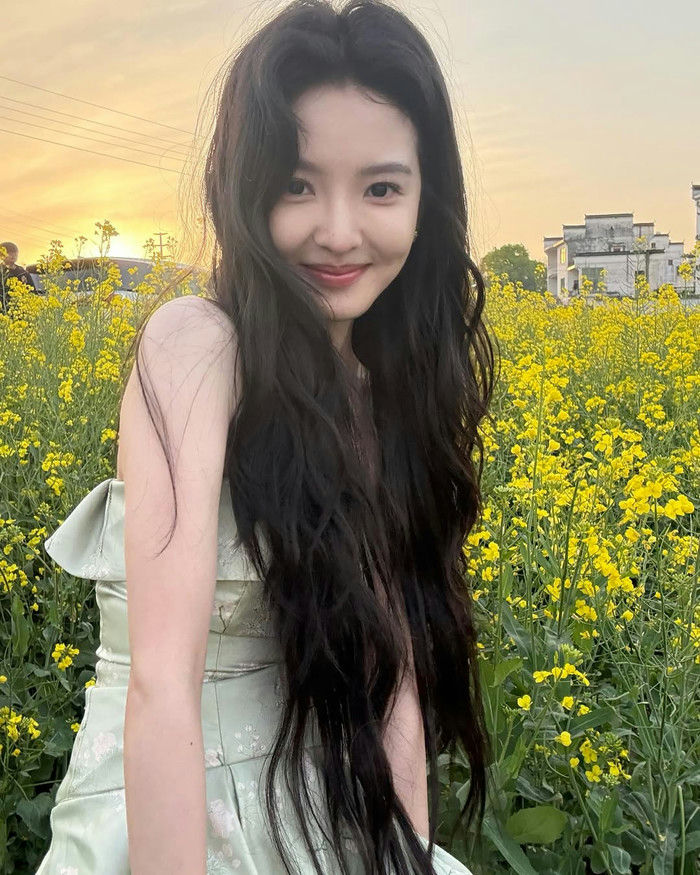 The photo features a young East Asian woman with long, dark, wavy hair, smiling gently at the camera. She is wearing a light mint green or sage green strapless dress with a subtle pattern. She is standing outdoors in a field densely populated with bright yellow flowers, likely rapeseed or canola blossoms, which dominate the background around her. The lighting suggests it is late afternoon or early evening, as the sky in the upper left corner is a warm golden hue from the setting or rising sun, with some utility lines visible. In the distance to the right, there are some simple, light-colored buildings. The person in the photo is Liu Yifei (Chinese actress). people-Liu Yifei-field of yellow flowers-smiling-long dark hair-green dress