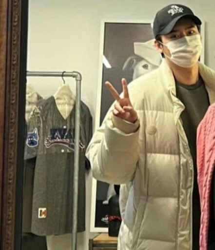 people-Man-wearing-a-mask-and-a-baseball-cap-making-a-peace-sign-while-wearing-a-light-colored-puffer-jacket-standing-in-front-of-a-clothing-rack-and-a-framed-picture
