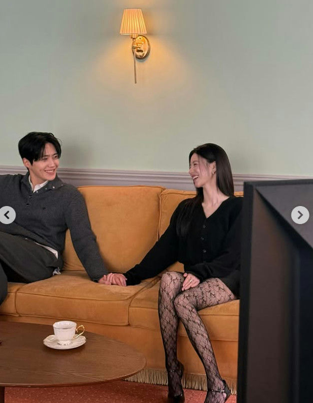 people-Kim Seon-ho-A-woman-in-black-cardigan-and-lace-stockings-A-sitting-on-mustard-couch-A-holding-hands-A-smiling-at-each-other-A-wall-sconce-light-fixture