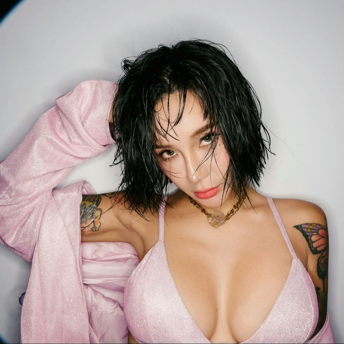 The photo features a woman with short, wet-look black hair, looking intensely at the camera. She is wearing a light pink, shimmery bikini top and is draped in a matching pink, slightly shiny jacket or robe pulled over one shoulder. She has visible tattoos on both arms, including what appears to be a butterfly wing on her right shoulder. She is also wearing a gold-toned chain necklace with a translucent pendant. The background is a plain, light color, possibly white or light gray. The person in the photo is Jessie J (or perhaps a model resembling her style, but this strongly appears to be the singer Jessie J). people-Jessie J-wet look hair-pink outfit-tattoos-bikini top-intense gaze