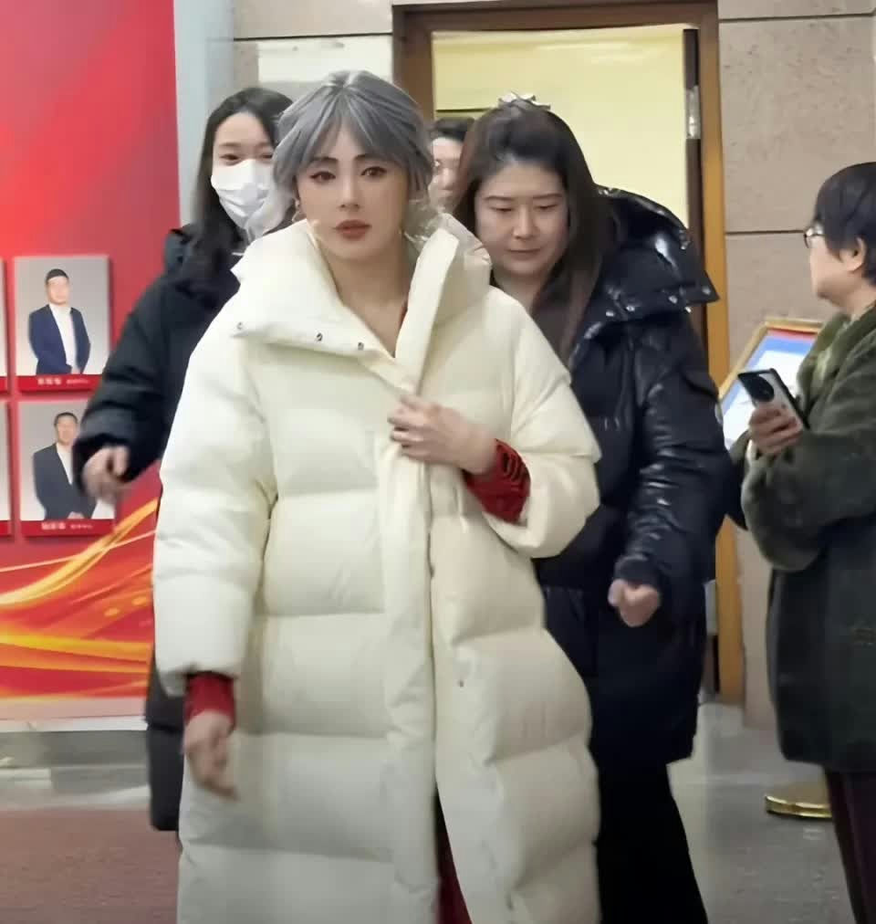 people-Woman with silver/grey hair and white puffer coat-Is the main focus-Woman wearing a white long puffer coat-Appears to be adjusting her coat-Woman to the right of the main subject-Wearing a black shiny puffer jacket-Woman to the far left-Wearing a black jacket and white face mask