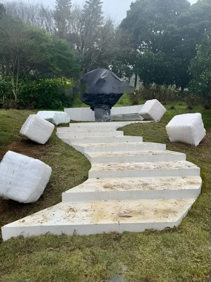 The image displays an outdoor scene, likely a garden or park under overcast or foggy conditions, featuring a set of newly constructed, curving white concrete steps ascending a grassy slope. At the top of the steps, there is a dark-colored sculpture or monument mounted on a pedestal, which is currently covered with a black protective tarp. Scattered around the base of the steps and the area near the monument are several large, roughly cube-shaped objects wrapped in white plastic or netting, resembling bales of material (perhaps insulation or raw material for construction/art). The background consists of dense green foliage and trees, some appearing somewhat obscured by mist or fog. No recognizable person appears in the photo. people-none-A-outdoor installation-A-sculpture under cover-B-curving steps-C-wrapped bales