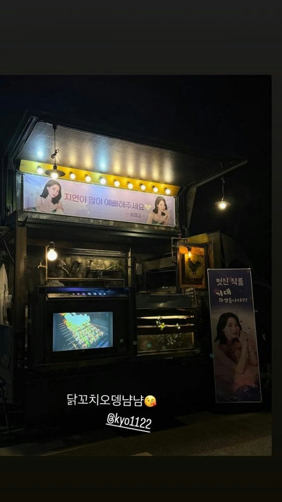This is a nighttime photo of a food truck, likely a catering or delivery setup for an event, illuminated by several lights. There are banners displayed prominently. The main banner features two images of a woman and Korean text that reads:
