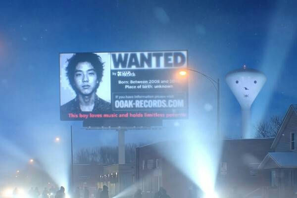 A nighttime, moody scene featuring a large, illuminated digital billboard. The billboard displays a black and white "WANTED" poster with the picture of a young East Asian male with dark, messy hair. Text on the billboard reads, in part: "by [logo] Records," "Born: Between 2008 and 20[text obscured]," "Place of birth: unknown," "If you have information please visit 00AK-RECORDS.COM," and "This boy loves music and holds limitless potential." Below the billboard, there are bright beams of light suggesting car headlights or stage lighting, and silhouettes of people on a street. To the right of the billboard, a distinctive water tower is visible, painted with a simple, cartoonish face (eyes, nose, and mouth indicated). The setting appears to be a suburban or urban area at night, possibly from a movie or promotional material. The person appearing in the photo is **T.O.P (Choi Seung-hyun)** from the K-pop group **Big Bang**, specifically from the promotional material for the film *Big Bang Made*. people-T.O.P (Choi Seung-hyun)-A-Wanted Billboard-A-Water Tower Face-B-Night Scene-C-K-pop Promotion