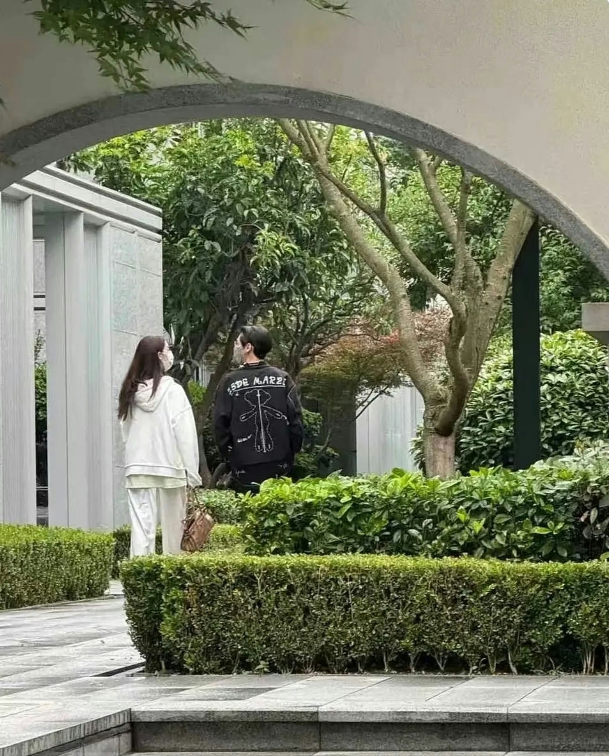 people-A-couple walking-A-park setting-A-man wearing black jacket with white graphic-A-woman in white loungewear-A-viewed through an archway