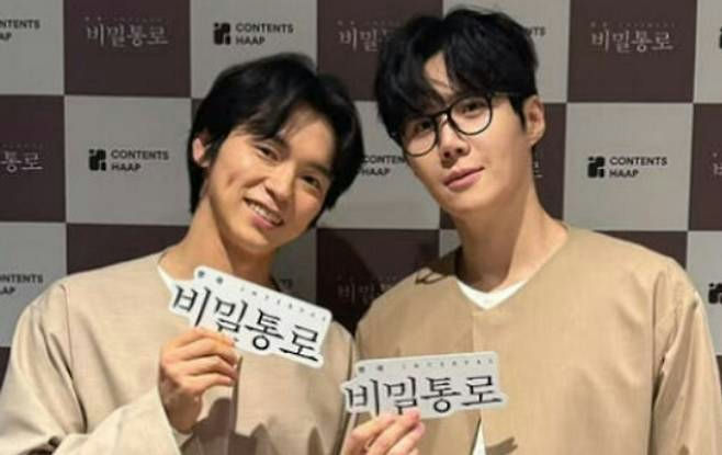 people-Two Korean men-A-holding signs-A-smiling-B-wearing glasses-C-background features checkered pattern and 