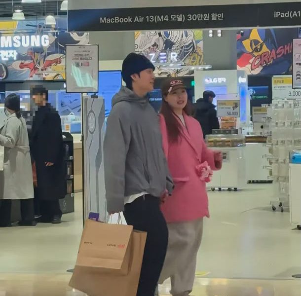 people-A-couple shopping-A-electronics store-B-man in grey hoodie and black beanie carrying shopping bags-C-woman in pink coat and cap-D-Korean text on banner advertising Apple products