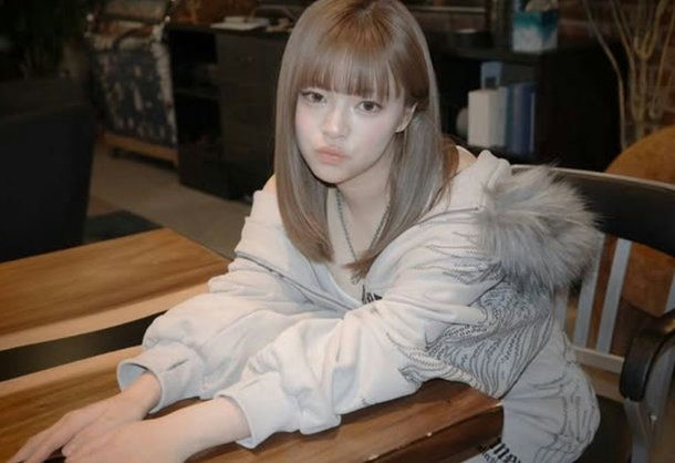 A young East Asian woman with light brown/ash-blonde hair and bangs is seated at a wooden table, leaning forward with her forearms resting on the table. She is wearing a light-colored (possibly white or very light gray) zip-up hoodie or jacket, which appears to have a fur-lined hood (the fur is light gray/white). The jacket has some subtle gray graphic print on the sleeves and body. She has a light complexion and is looking directly at the camera with a slightly pouty or neutral expression. The background is somewhat dimly lit, suggesting an indoor setting like a cafe or lounge, with dark furniture and some blurred objects visible behind her. **Person Identification:** Based on visual appearance, this individual is **Yoo Jeongyeon** (Jeongyeon) from the K-pop group TWICE. people-Yoo Jeongyeon-East Asian woman-ash blonde hair-fur-lined hoodie-pouty expression-indoor setting