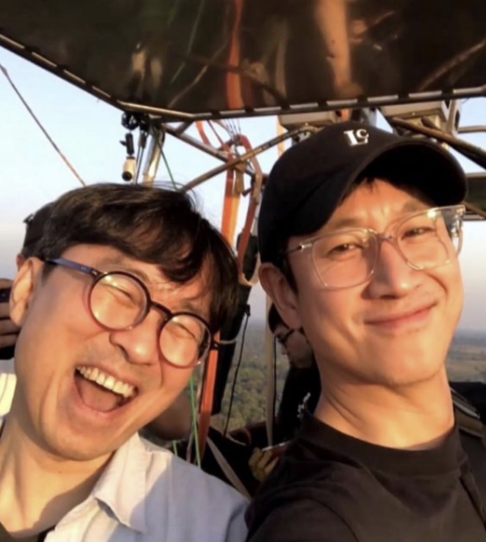 people-Two Asian men-are smiling-while riding in a hot air balloon basket-They appear to be traveling or on vacation-The man on the left is laughing heartily with round glasses and a light-colored shirt, and the man on the right is wearing a black cap and clear-framed glasses-The photo is a selfie taken outdoors, likely during sunrise or sunset, given the warm light.
